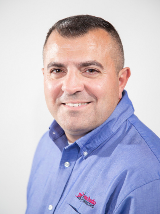 A headshot of Helder in a blue collared shirt, smiling at the camera. He has short brown hair and dark eyebrows. The Machado logo is on his shirt 