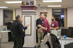 5 people are standing around a column in an office chatting. Two are in maroon shirts and three are wearing black.