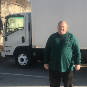 A bald man in a green shirt stands ouside of a large white delivery truck. 