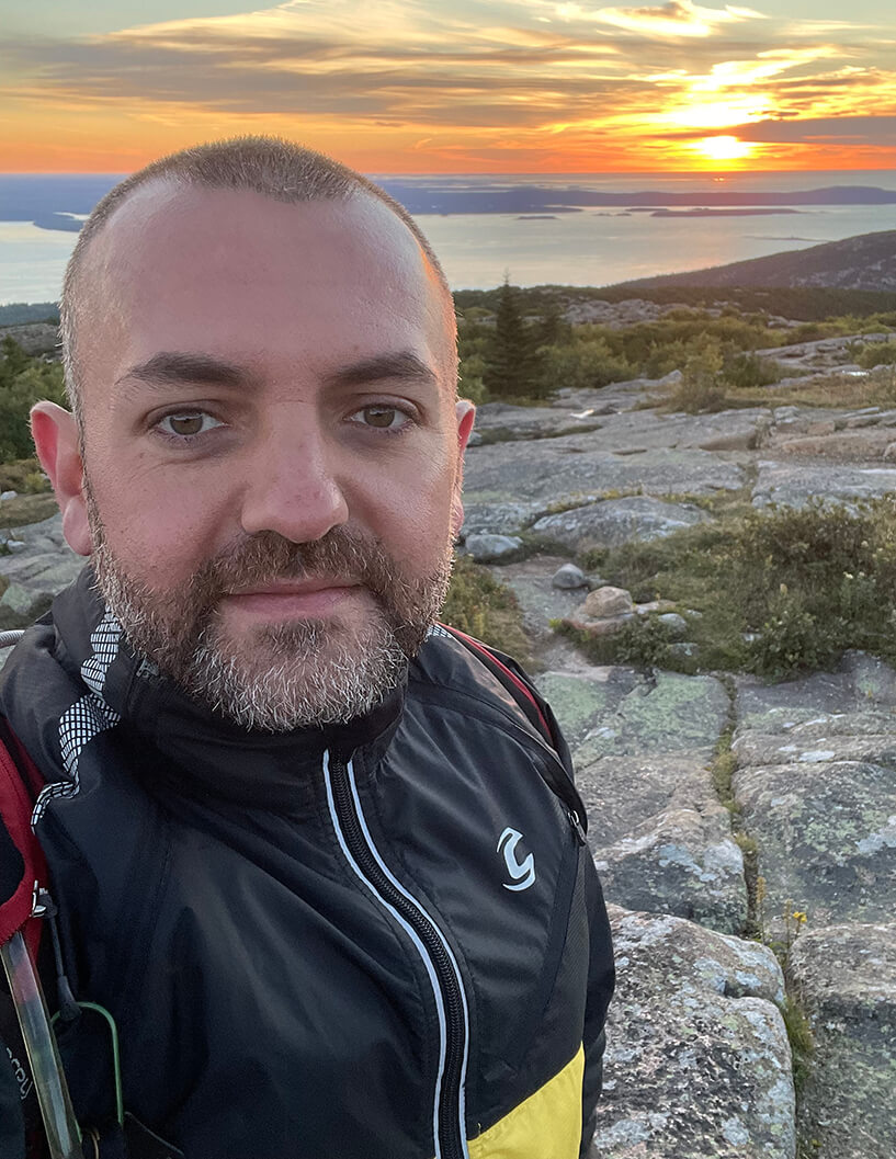 A man with a buzzcut slightly smiles at the camera with a sunset in the background at the top of a hike. 