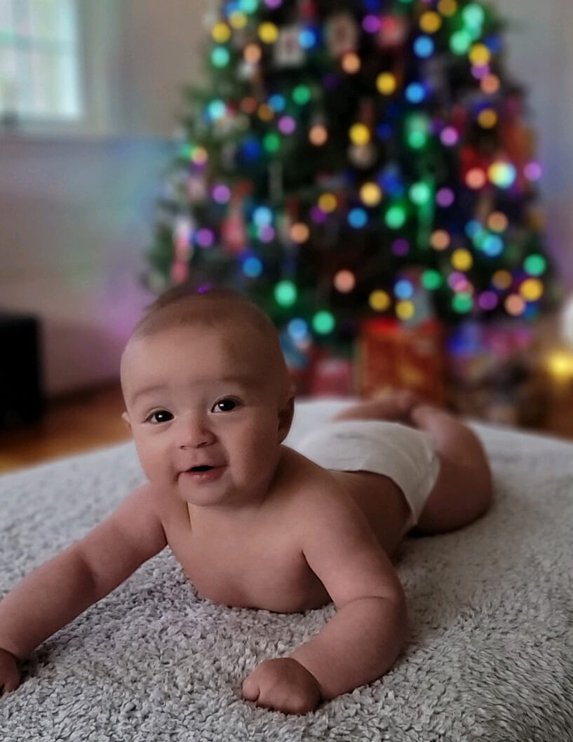 A baby smiling at the camera, laying on their stomach with a christmas tree blurred in the background.