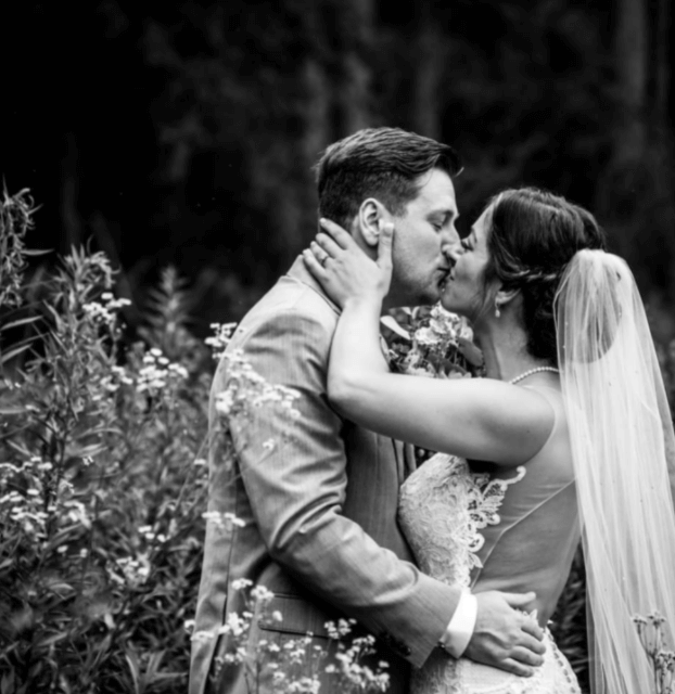 A man and woman kissing on their wedding day, surrounded by large plants. All in black and white. 